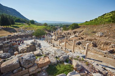 Antik Yunan kenti Efes 'teki Celsus Kütüphanesi' ne uzanan sütunları olan mermer bir ara sokakta dağlar ve arka planda açık mavi gökyüzü, Türkiye 'nin Celsus Kütüphanesi üzerinde tek bir küçük bulut var.