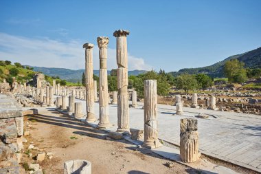 Antik Yunan kenti Efes 'teki Celsus Kütüphanesi' ne uzanan sütunları olan mermer bir ara sokakta dağlar ve arka planda açık mavi gökyüzü, Türkiye 'nin Celsus Kütüphanesi üzerinde tek bir küçük bulut var.