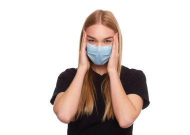 Kadın virüse karşı koruyucu beyaz bir maske takıyor, şakaklarında şiddetli bir acı hissediyor, parmaklarını kafaya doğru tutuyor. Siyah tişörtlü, uzun boylu sarışın bir kız. Stüdyoda beyaz arka planda izole edilmiş.