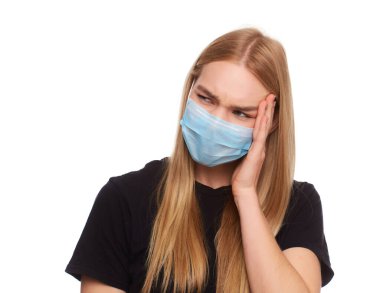Kadın virüse karşı koruyucu beyaz bir maske takıyor, şakaklarında şiddetli bir acı hissediyor, parmaklarını kafaya doğru tutuyor. Siyah tişörtlü, uzun boylu sarışın bir kız. Stüdyoda beyaz arka planda izole edilmiş.