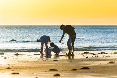 Çocuklar plajda balıkçı ağıyla oynuyorlar. Gün batımı deniz manzarası, gün batımına karşı insan silueti.
