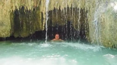 Mavi suyla dolu bir gölde şelalenin altında yüzen kızıl saçlı güzel bir kadın. 4K görüntü klibi