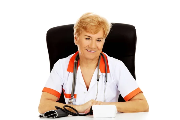 Sorria idosa médica ou enfermeira sentada atrás da mesa com medidor de preasure bloog — Fotografia de Stock