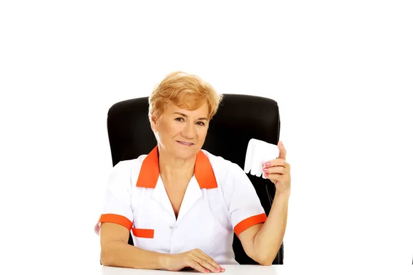 Smile elderly female dentist sitting behind the desk and holds big tooth model — Stock Photo, Image
