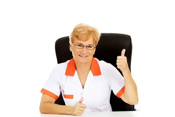 微笑老年女性医生或护士坐在桌子和节目拇指后面 — 图库照片