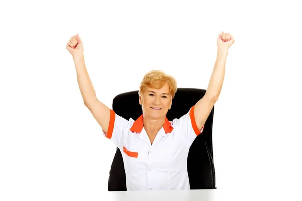 Feliz anciana doctora o enfermera sentada detrás del escritorio con las manos arriba —  Fotos de Stock