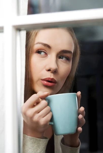 Belle femme caucasienne buvant du café chaud ou du thé et regardant par la fenêtre . — Photo