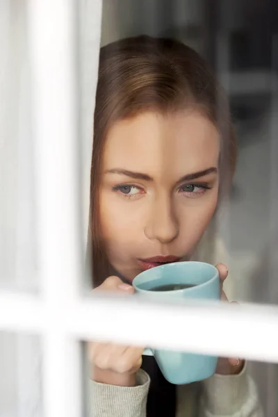 Bella donna caucasica che beve caffè caldo o tè e guarda attraverso la finestra . — Foto Stock