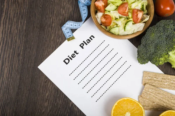 Food and sheet of paper with a diet plan on a dark wooden table. — Stock Photo, Image