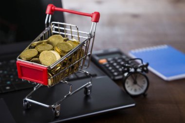 Bir tablo üzerinde paralar, dizüstü bilgisayar ve çalar saat ile alışveriş arabası.