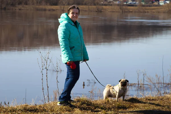 Woman with a dog walks on the riverbank on a spring day. Leisure.