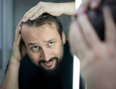 Aynanın önündeki adam saçlarını ve tenini kontrol ediyor.