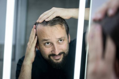 Aynanın önündeki adam saçlarını ve tenini kontrol ediyor.
