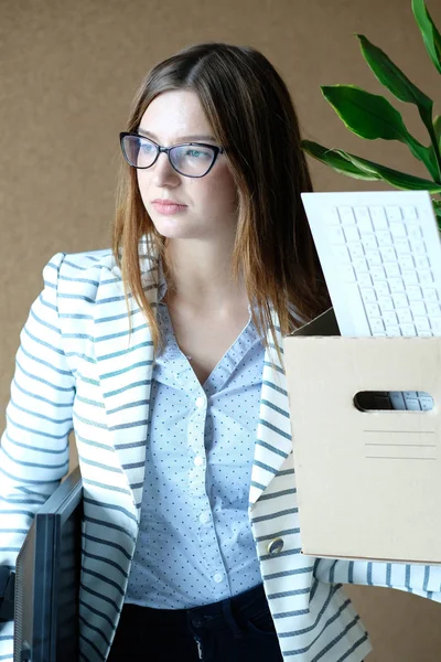 young woman being fired from work, holding box with pot plant and personal stuff