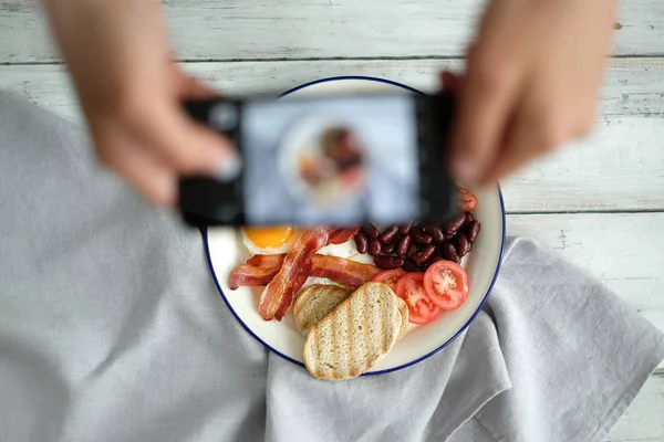 man taking photo of English breakfast with smartphone: egg, bacon, sausages, mushrooms, tomatoes, beans and bread