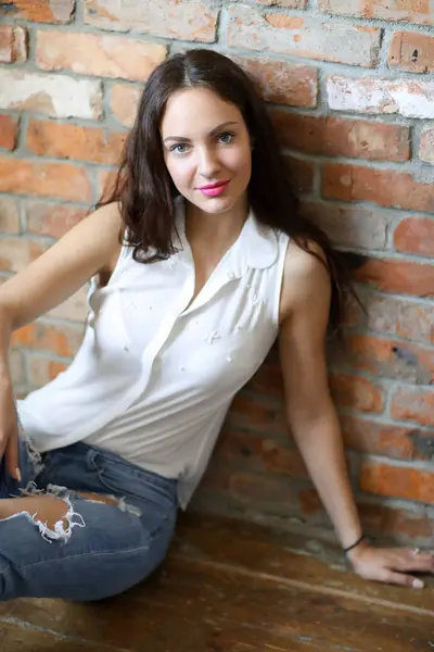 young brunette woman in jeans leaning against brickwall at home sitting on floor