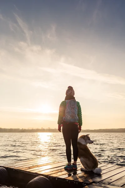 Femme avec chien profiter du lever du soleil au lac, silhouette sac à dos — Photo