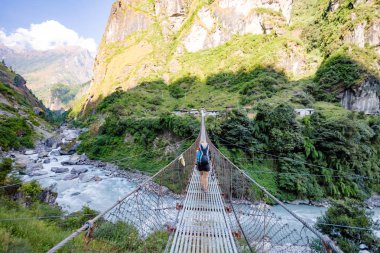 Woman backpacker crossing suspension bridge in Himalayas Nepal clipart