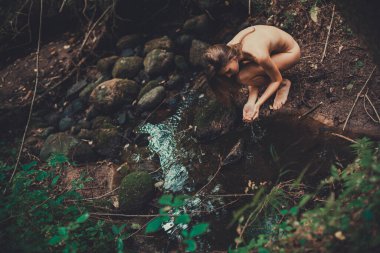 Nehrin kıyısında oturmuş su içen çıplak bir kadının fotoğrafı.