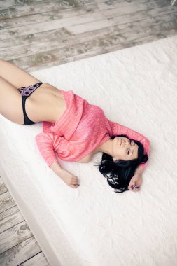 Pembe kazaklı, siyah külotlu güzel gülüşlü esmer kız beyaz bir yatakta uzanıyor ve mükemmel vücudunu gösteriyor.