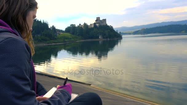 Chica dibujando un boceto de un castillo Niedzica sentado en una orilla del lago — Vídeo de stock