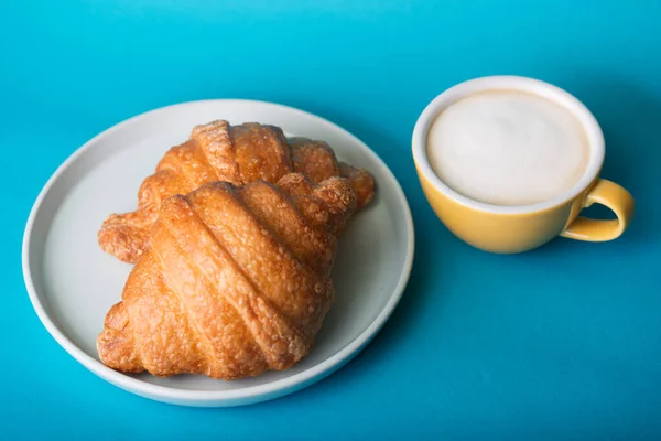 Cangkir kuning dengan croissant pada latar belakang biru — Stok Foto