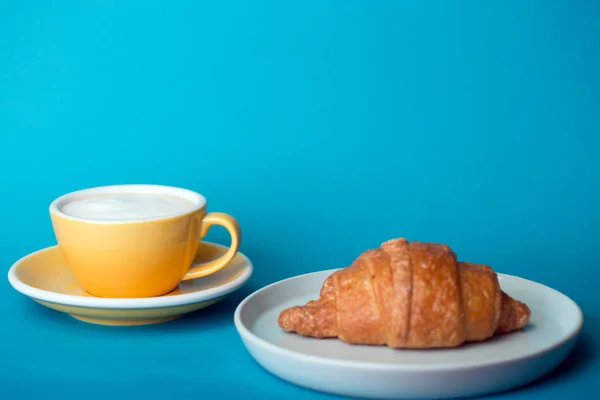Gelbe Tasse mit Croissant auf blauem Hintergrund — Stockfoto