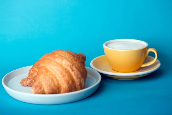 Gelbe Tasse mit Croissant auf blauem Hintergrund — Stockfoto