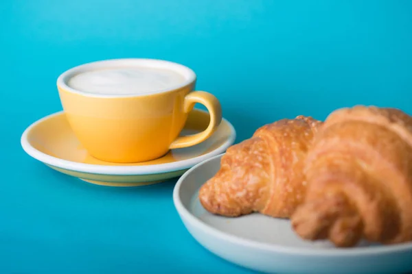 Gelbe Tasse mit Croissant auf blauem Hintergrund — Stockfoto