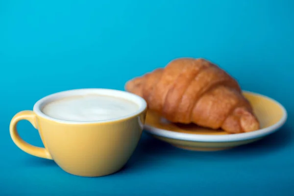 Gelbe Tasse mit Croissant auf blauem Hintergrund — Stockfoto