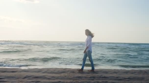 夏天刮风的日子里 身穿牛仔裤和白衬衫的女孩在海滨带着大浪散步 — 图库视频影像