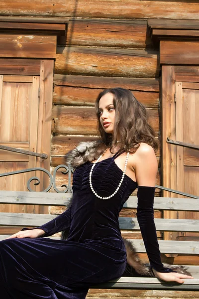 Young girl in an evening dress on a background of the old wooden houses with shutters closed — Stock Photo, Image