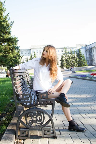 Chica en camisa blanca y pantalones cortos sentados en un banco en el parque — Foto de Stock