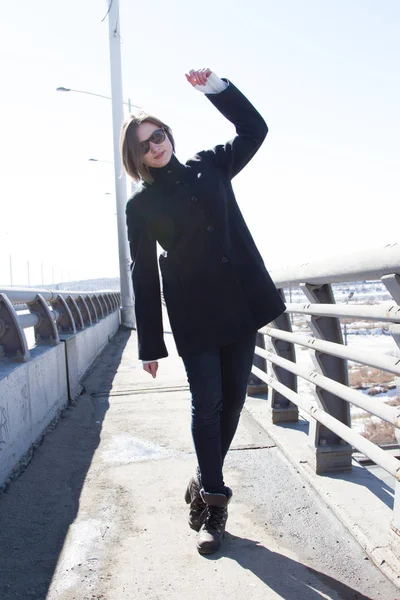 Young girl in a coat and jeans is standing on a bridge — Stock Photo, Image