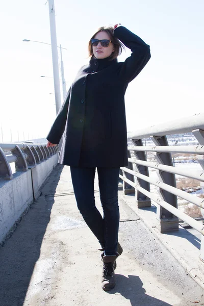 Young girl in a coat and jeans is standing on a bridge — Stock Photo, Image