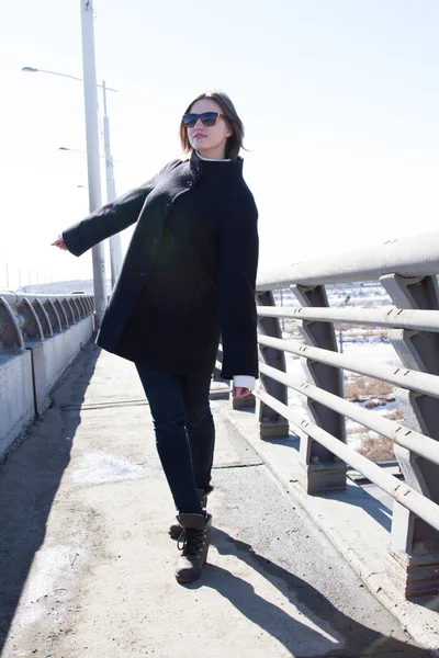 Joven chica en un abrigo y jeans está de pie en un puente —  Fotos de Stock