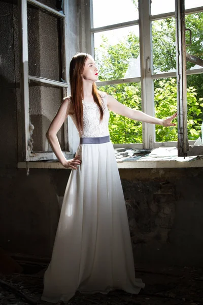 Mädchen in weißem Kleid steht im Hintergrund eines Fensters in einem verlassenen Haus — Stockfoto