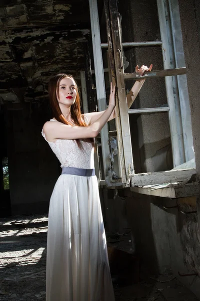 Mädchen in weißem Kleid steht im Hintergrund eines Fensters in einem verlassenen Haus — Stockfoto