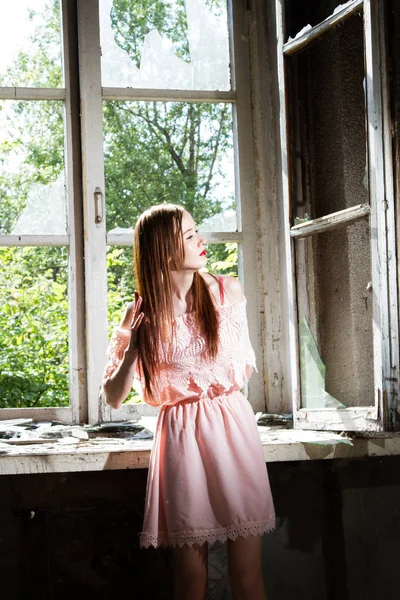 Menina em um vestido curto rosa está de pé pela janela em uma casa abandonada — Fotografia de Stock