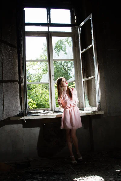 Chica en un vestido corto de color rosa está de pie junto a la ventana en una casa abandonada — Foto de Stock