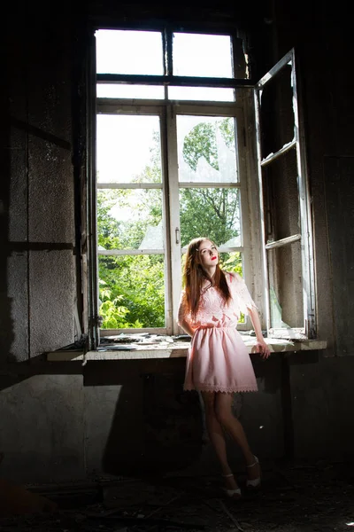 Chica en un vestido corto de color rosa está de pie junto a la ventana en una casa abandonada — Foto de Stock