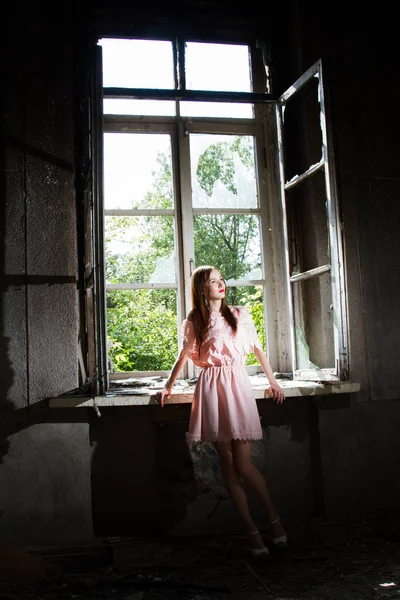 Mädchen in kurzem rosa Kleid steht am Fenster in einem verlassenen Haus — Stockfoto