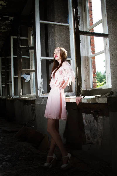 Mädchen in kurzem rosa Kleid steht am Fenster in einem verlassenen Haus — Stockfoto