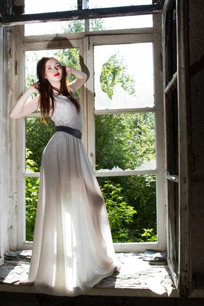 Schlankes junges Mädchen in weißem Kleid auf Fensterbank in verlassenem Haus — Stockfoto