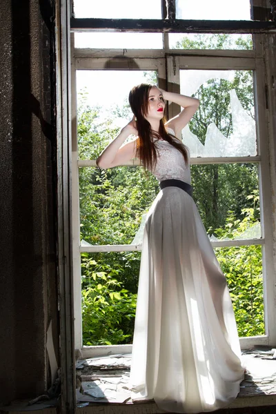 Стройная молодая девушка в белом платье на подоконнике в заброшенном доме — стоковое фото