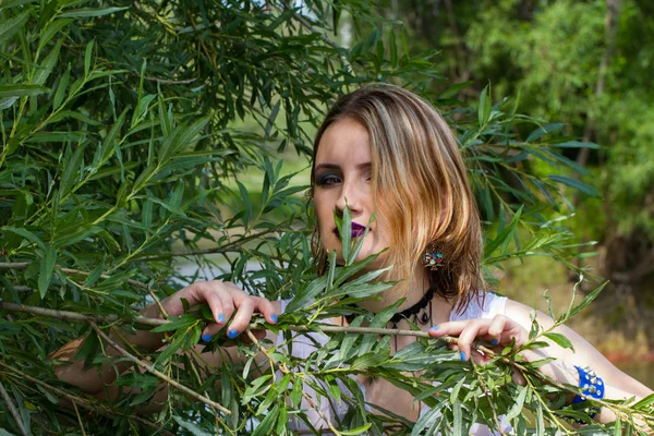Girl with bright makeup looks out from behind branches — Stock Photo, Image