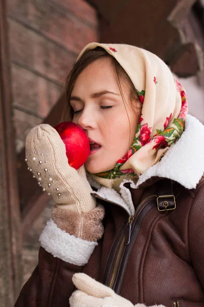 Girl in a colorful scarf and with a red apple is standing on the porch of an old wooden house — Stock Photo, Image