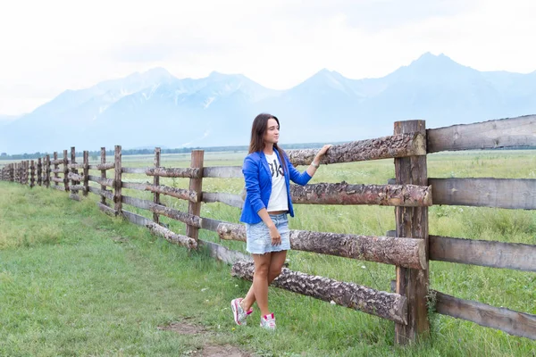 Νεαρό κορίτσι σε ένα μπλε σακάκι στέκεται σε ένα μακρύ ξύλινο φράχτη — Φωτογραφία Αρχείου