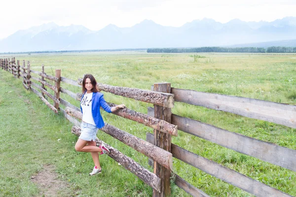 Νεαρό κορίτσι σε ένα μπλε σακάκι στέκεται σε ένα μακρύ ξύλινο φράχτη — Φωτογραφία Αρχείου