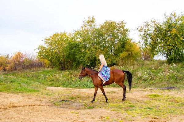 A girl in an evening dress jumps on a horse on the shore of a pond — Stock Photo, Image